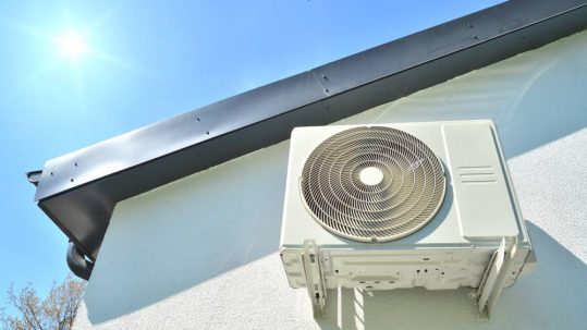 An air conditioning unit is installed to the side of an exterior wall of a home in the sun with a blue sky.