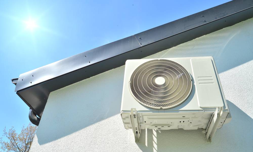 An air conditioning unit is installed to the side of an exterior wall of a home in the sun with a blue sky.