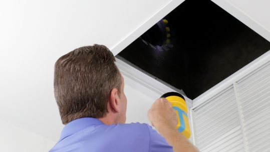 A man wearing a purple polo shirt and holding a yellow flashlight inspects a home's air duct with the air vent removed.