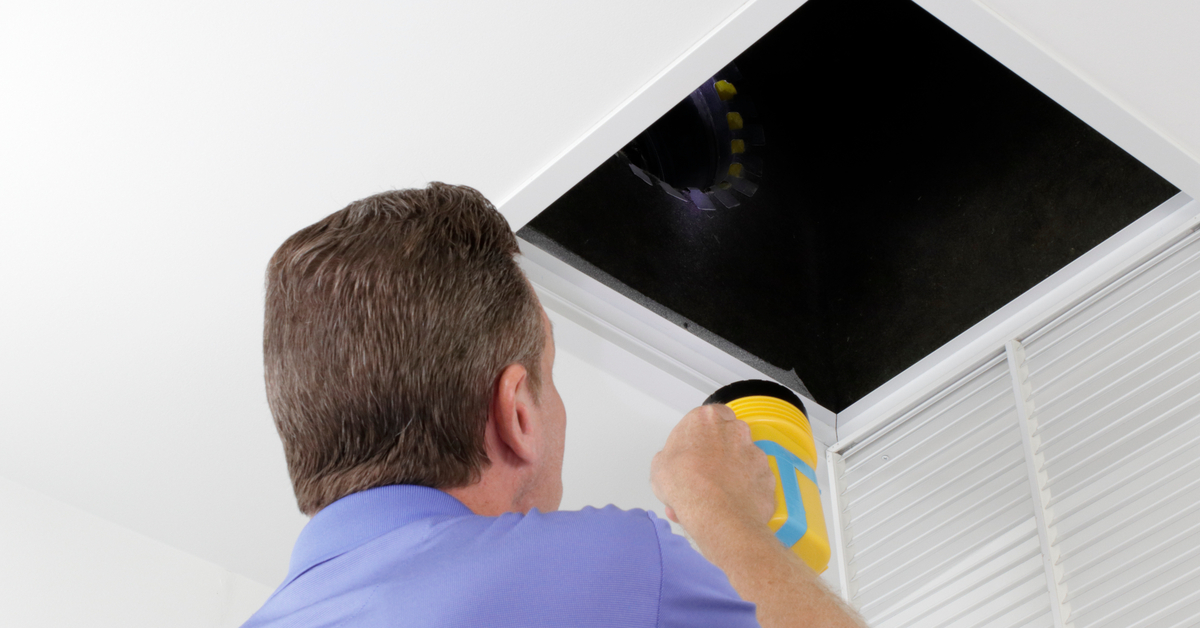 A man wearing a purple polo shirt and holding a yellow flashlight inspects a home's air duct with the air vent removed.