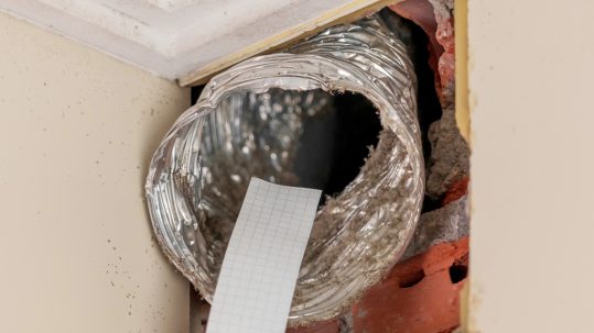 A technician's hand holds a strip of paper to an exposed air duct in a home to inspect its ventilation.