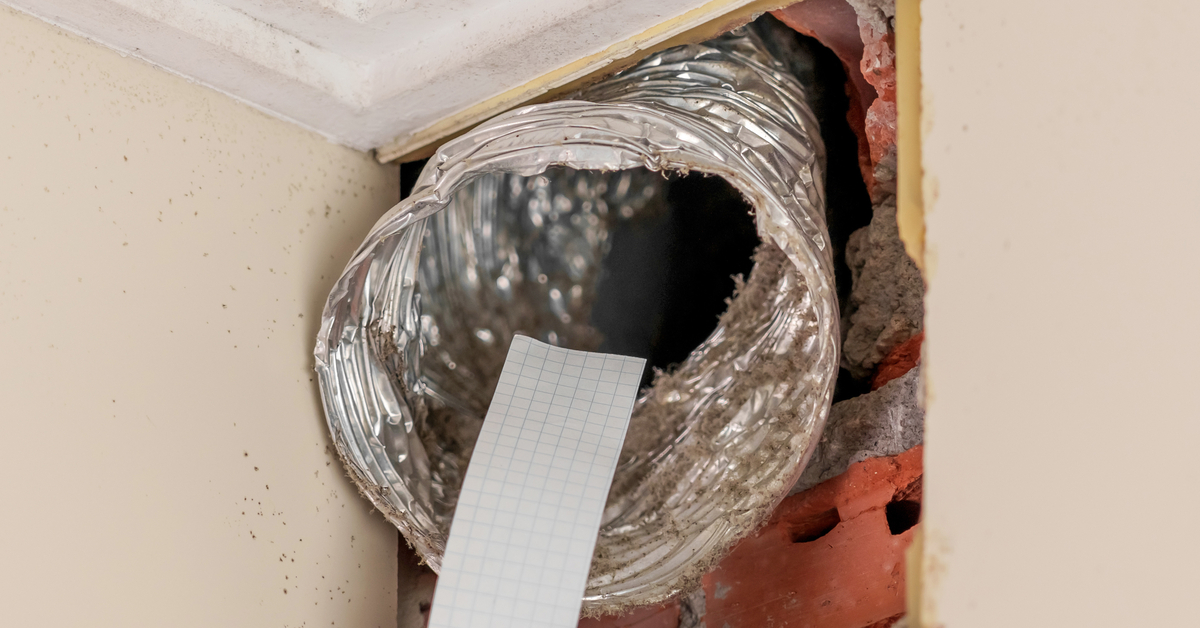 A technician's hand holds a strip of paper to an exposed air duct in a home to inspect its ventilation.