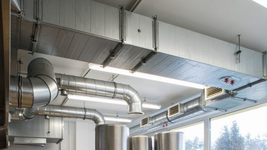 An exposed commercial air duct system in a kitchen interior. The ducts hang from the ceiling in between fluorescent lights.