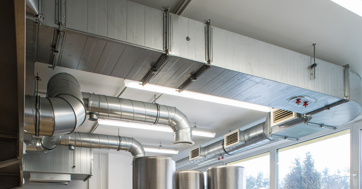 An exposed commercial air duct system in a kitchen interior. The ducts hang from the ceiling in between fluorescent lights.