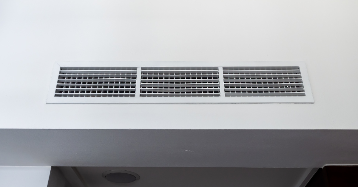 A long, rectangular air ventilation grill free of dust and debris on a plain white wall above a walkway.