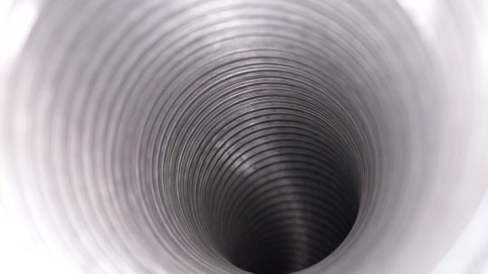 An up-close view of the inside of an aluminum air duct. The duct has ridges throughout and curves downward.