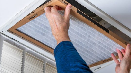 A pair of hands reaching up and replacing a dirty HVAC air filter in a ceiling vent. The person is wearing a blue shirt.