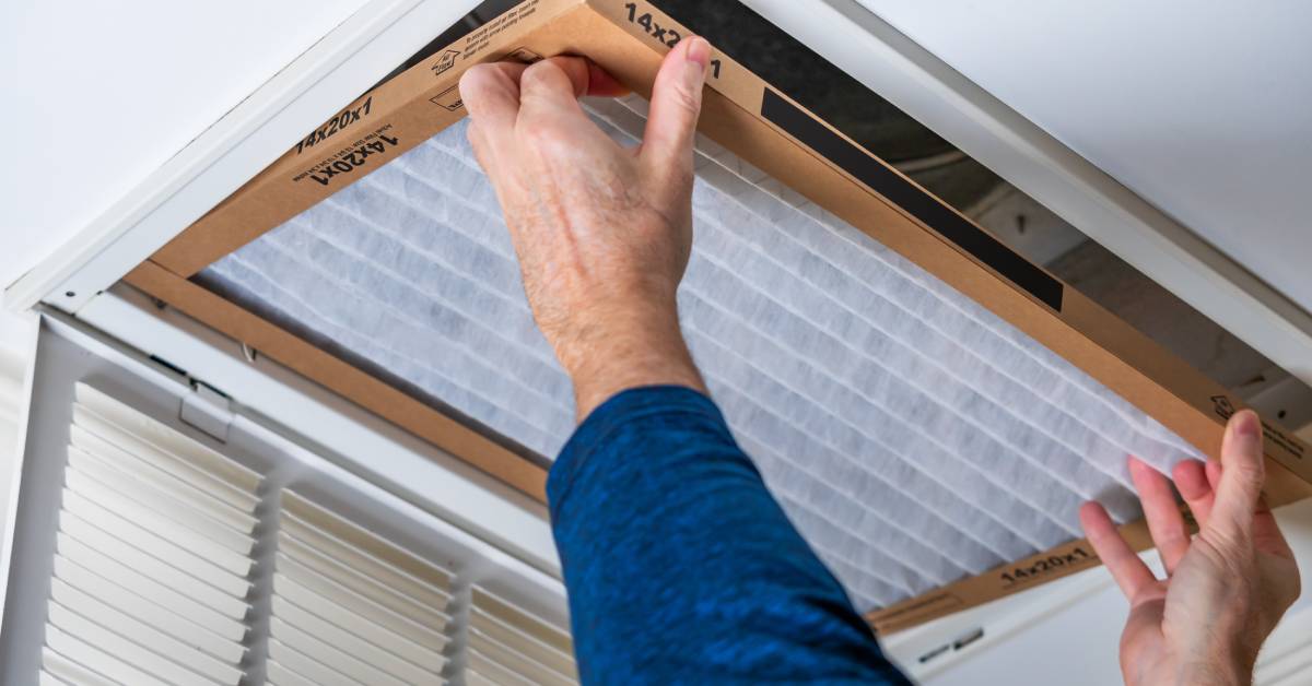 A pair of hands reaching up and replacing a dirty HVAC air filter in a ceiling vent. The person is wearing a blue shirt.